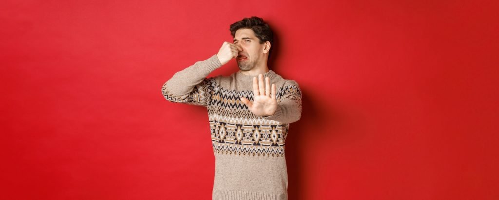 Image of disgusted handsome man in winter sweater, rejecting something with bad smell, shut nose
